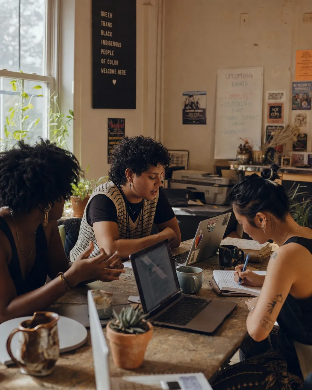 Three LGBTQ+ community members working together around a cluttered table in a casual community space, one mid-gesture in conversation, another pointing at a laptop screen.